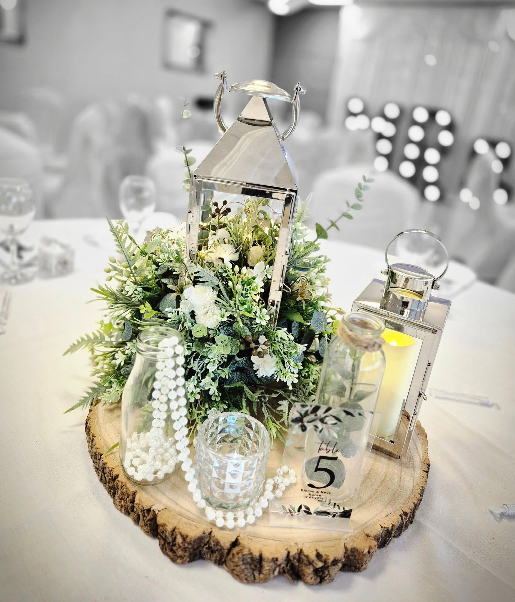 Wedding table centerpiece featuring silver lanterns, pearl accents, and greenery on rustic wood base