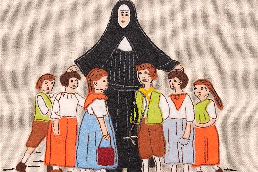 Gestickte Darstellung einer Nonne in schwarzer Ordenstracht, die schützend ihre Arme um sechs Kinder legt. Die Kinder tragen bunte Kleidung in Orange-, Blau- und Brauntönen. Alle Figuren stehen auf hellem Stoffhintergrund.