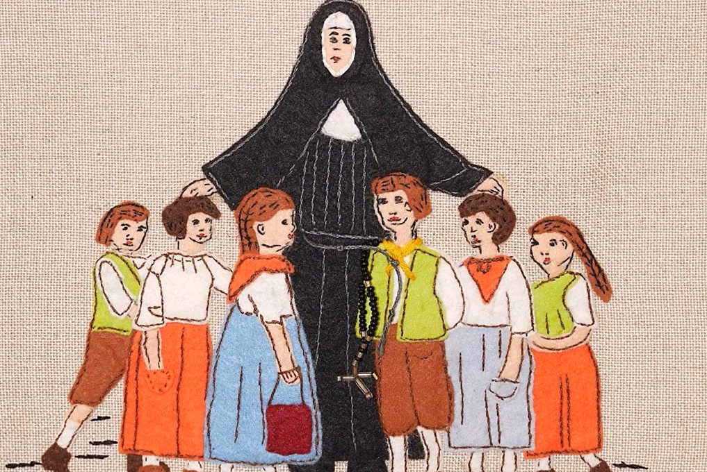 Gestickte Darstellung einer Nonne in schwarzer Ordenstracht, die schützend ihre Arme um sechs Kinder legt. Die Kinder tragen bunte Kleidung in Orange-, Blau- und Brauntönen. Alle Figuren stehen auf hellem Stoffhintergrund.
