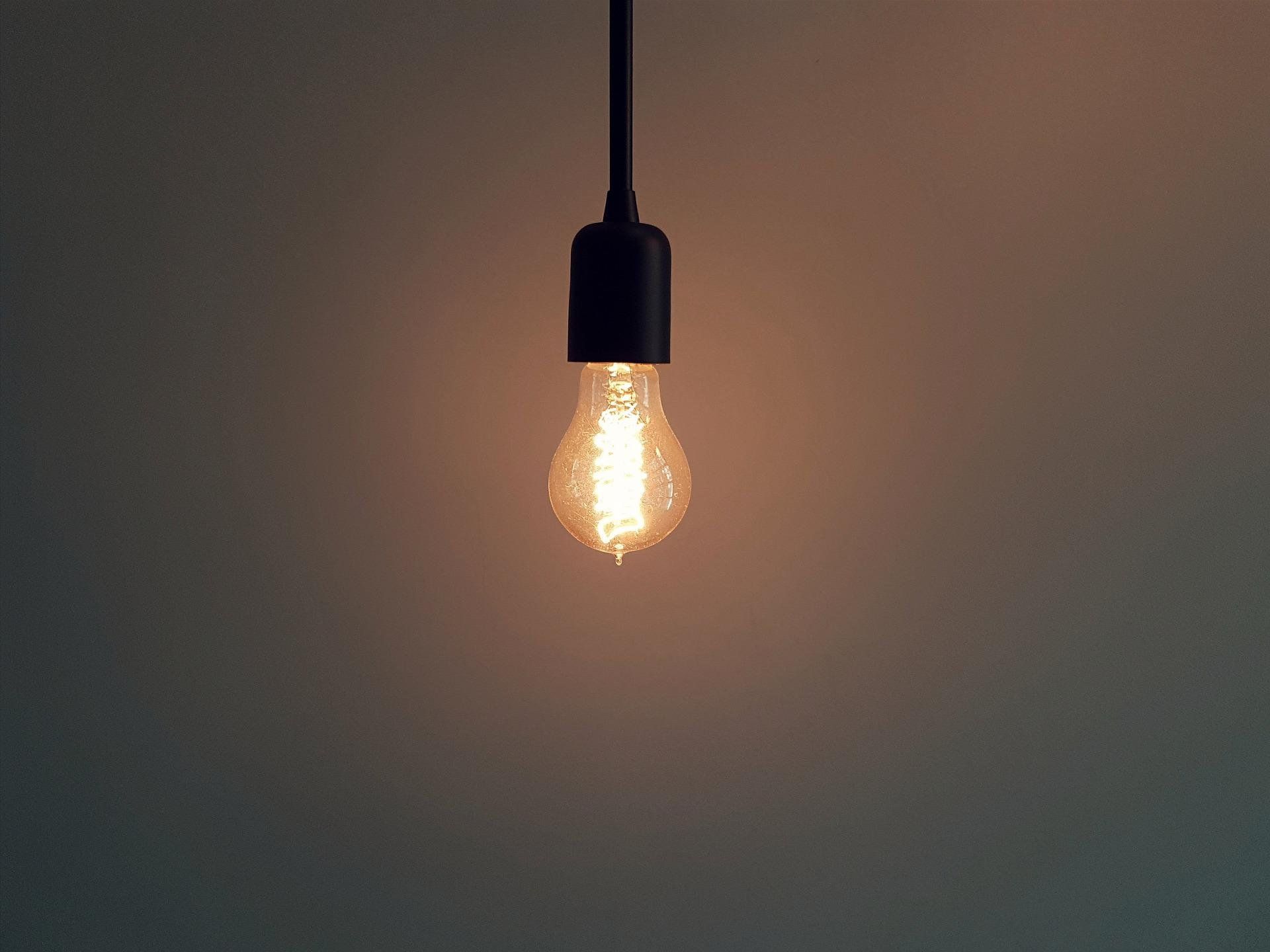 Glühbirne hängt an Kabel vor dunklem Hintergrund, der etwas beleuchtet wird