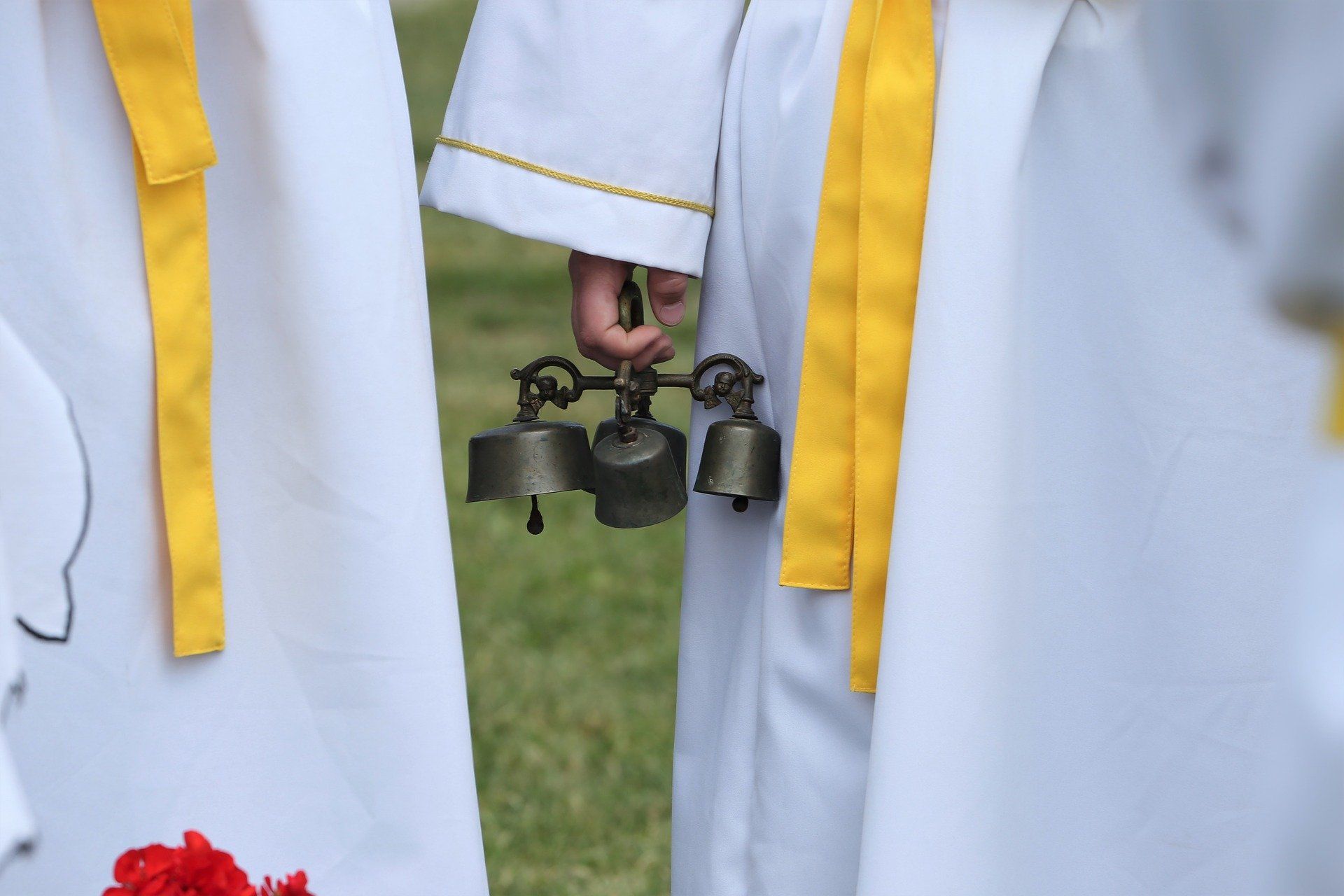 Zwei Personen in weißen liturgischen Gewändern mit gelben Bändern stehen nebeneinander. Eine Hand hält mehrere kleine Altarschellen. Im Vordergrund sind rote Blumen angeschnitten.