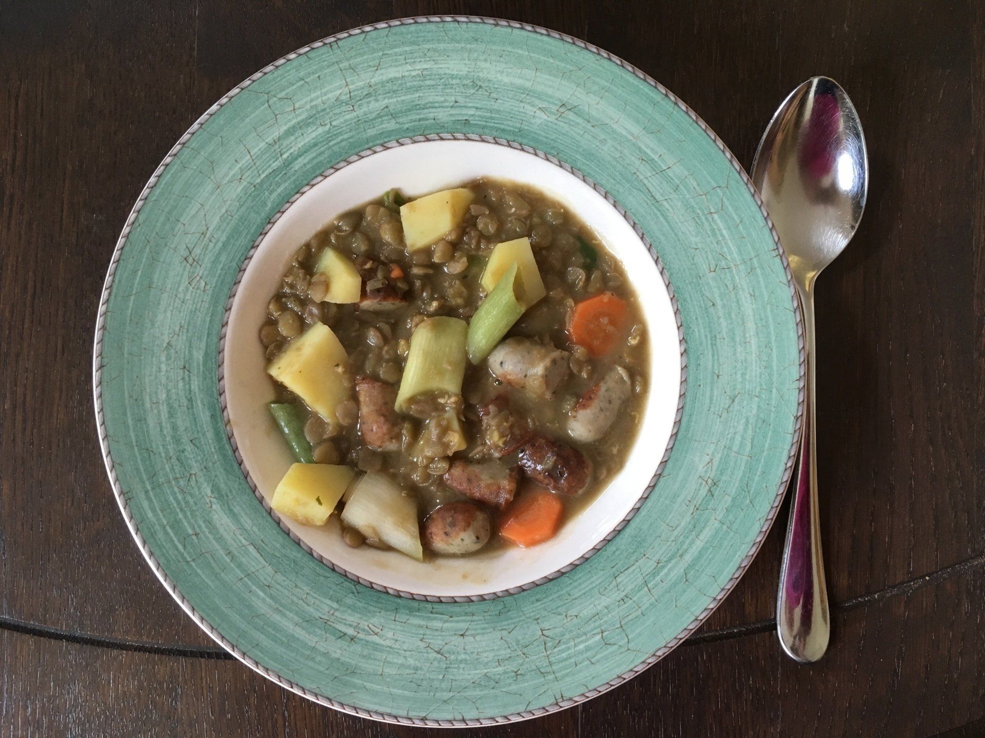 Ein tiefer Teller mit grünem Rand, gefüllt mit einem Linseneintopf. In der Suppe sind unter anderem Kartoffeln, Möhren, grüne Bohnen und Würstchenscheiben zu sehen. Rechts daneben liegt ein Löffel auf dem dunklen Holztisch.