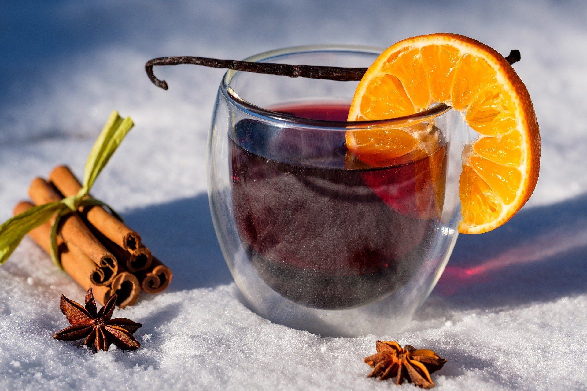Ein Glas mit rotem Tee steht im Schnee, verziert mit einer Orangenscheibe und einer Vanilleschote. Um die Tasse herum liegen Zimtstangen und Stern-Anis.