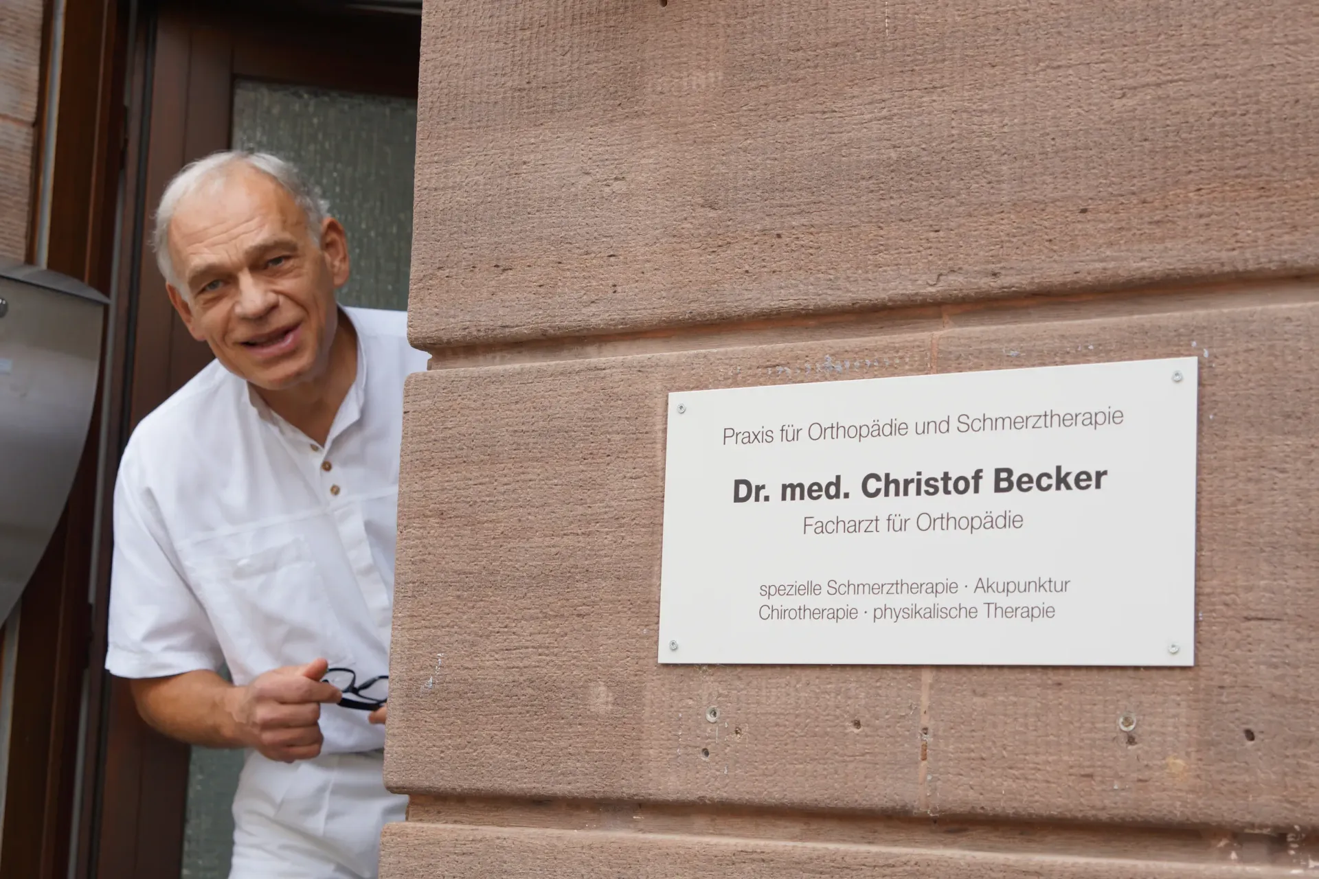Dr. Becker steht im Eingangsbereich der Praxis und schaut freundlich in die Kamera. Auf der Hauswand daneben ist das Adressenschild der Praxis befestigt