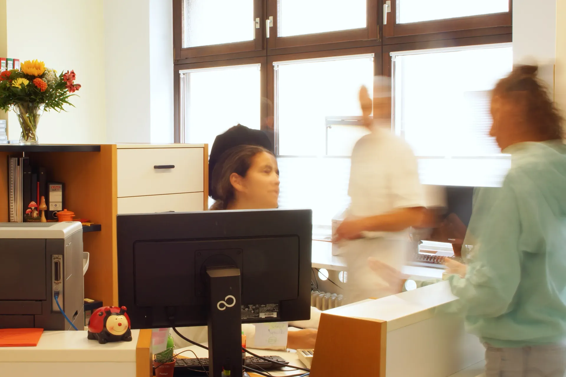 Eingangsbereich der Praxis. Ein Patient steht vor dem Empfangstresen. In unscharfer Bewegung dahinter Herr Dr. Becker und Personal