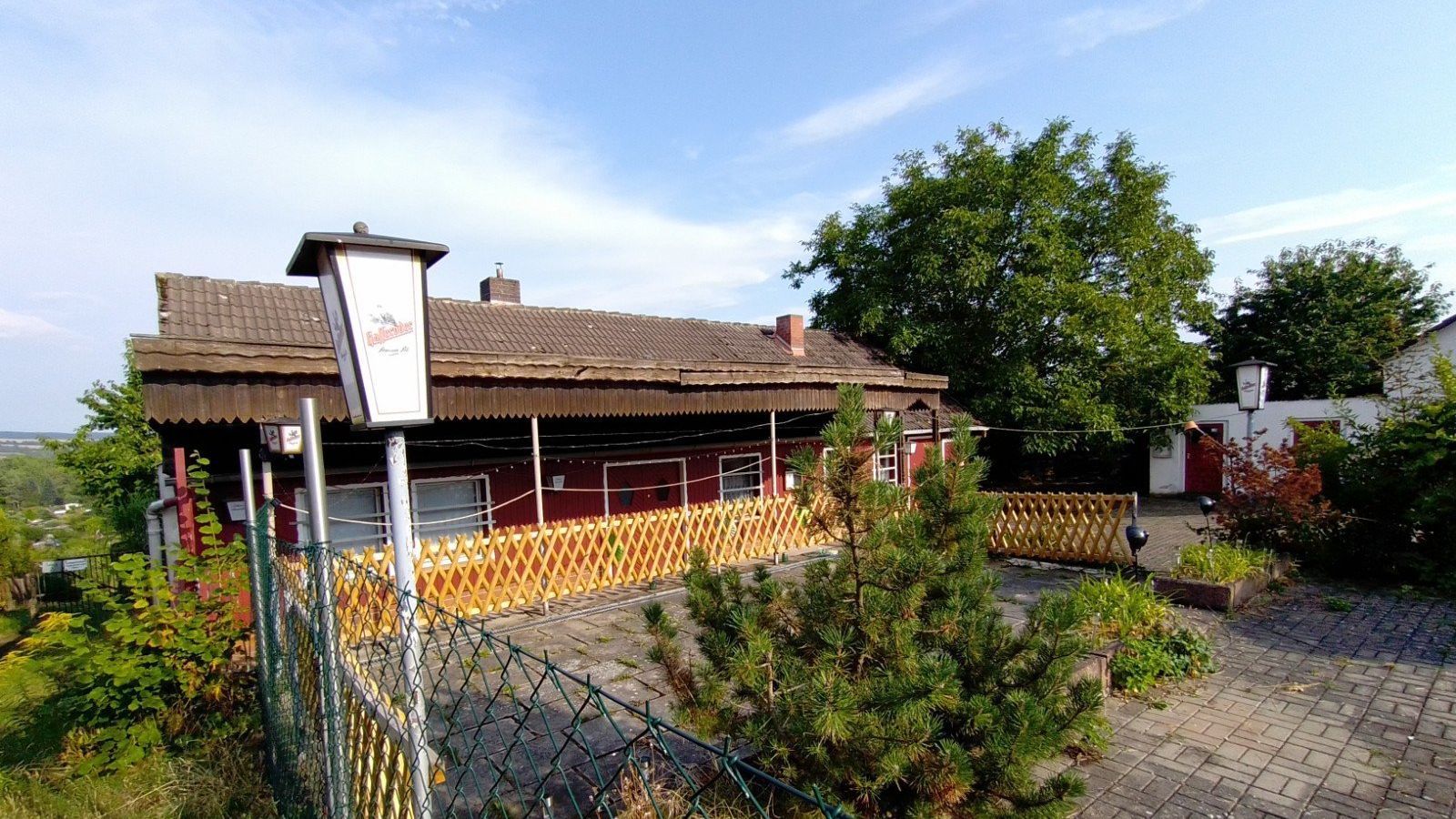 Das Vereinsheim des KGV ‚Schöne Aussicht‘ Nordhausen ist ein eingeschossiges, rot gestrichenes Gebäude mit überdachter Terrasse und gelbem Holzgeländer. Es liegt an einem gepflasterten Weg und ist von Bäumen, Sträuchern und kleinen Hinweisschildern umgeben. Die Aufnahme zeigt den ruhigen Außenbereich der Kleingartenanlage.