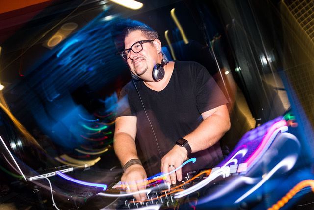 Hochzeits-DJ hat die Hände an seinem Pult und lächelt zu Publikum. Er trägt einen Brille, Bart und T-Shirt mit V-Ausschnitt.