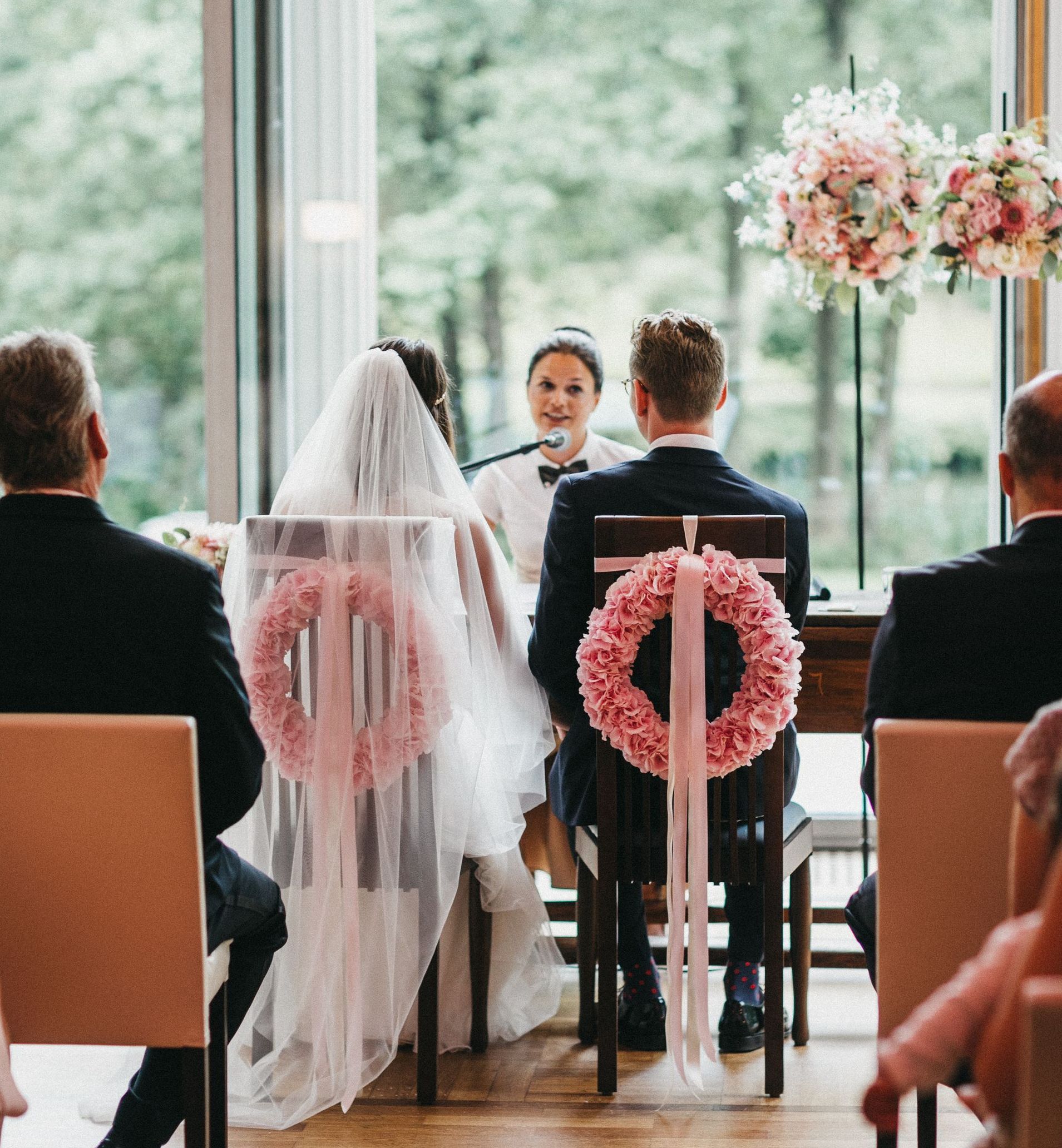 Zu sehen ist eine Freie Trauung in Hamburg. Das Brautpaar sitzt mit dem Rücken zu den Gästen auf Stühlen, die mit wunderschönen rosafarbenen Hortensienkränzen geschmückt sind.