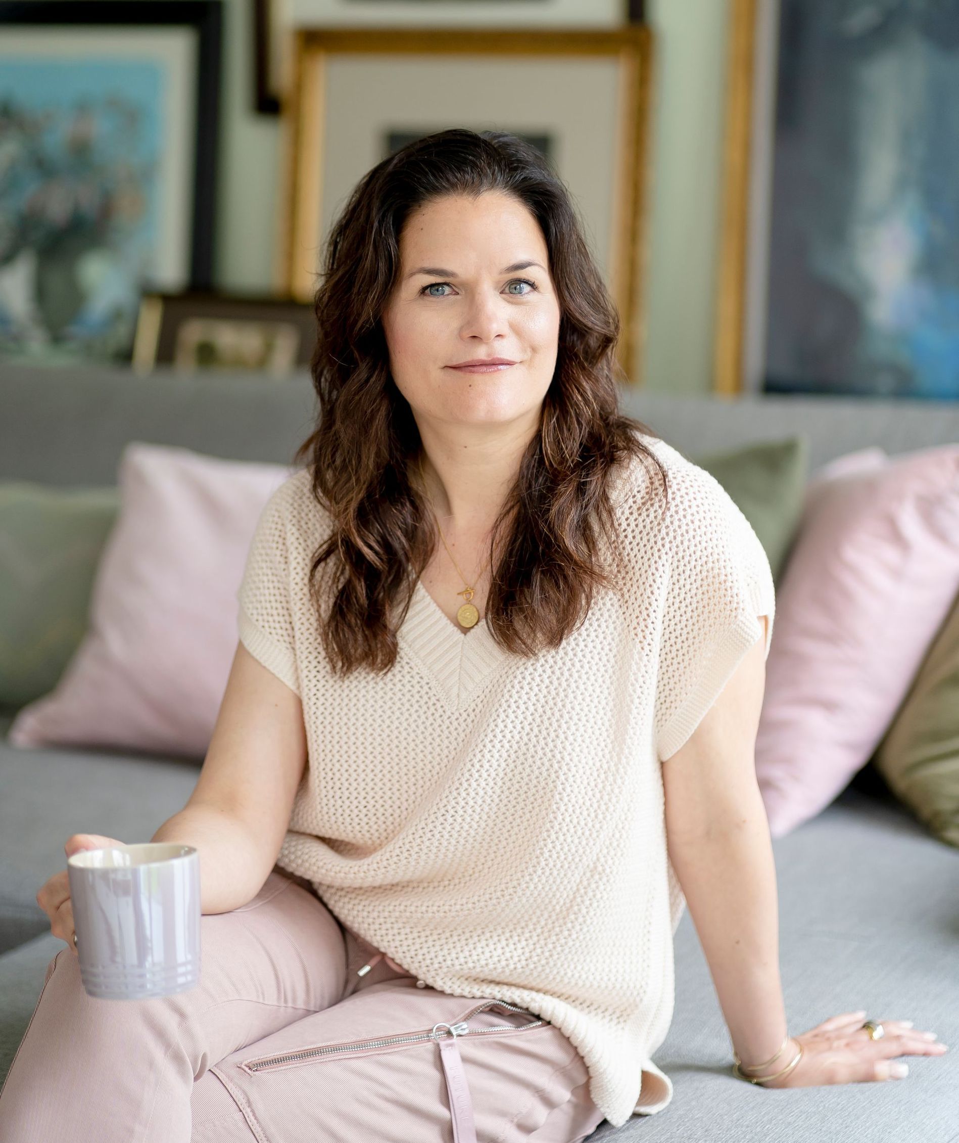 Freie Traurednerin Kristina Kutz sitzt auf einem hellgrauen Sofa inmitten von vielen Kissen. Sie hält eine Tasse in der Hand.