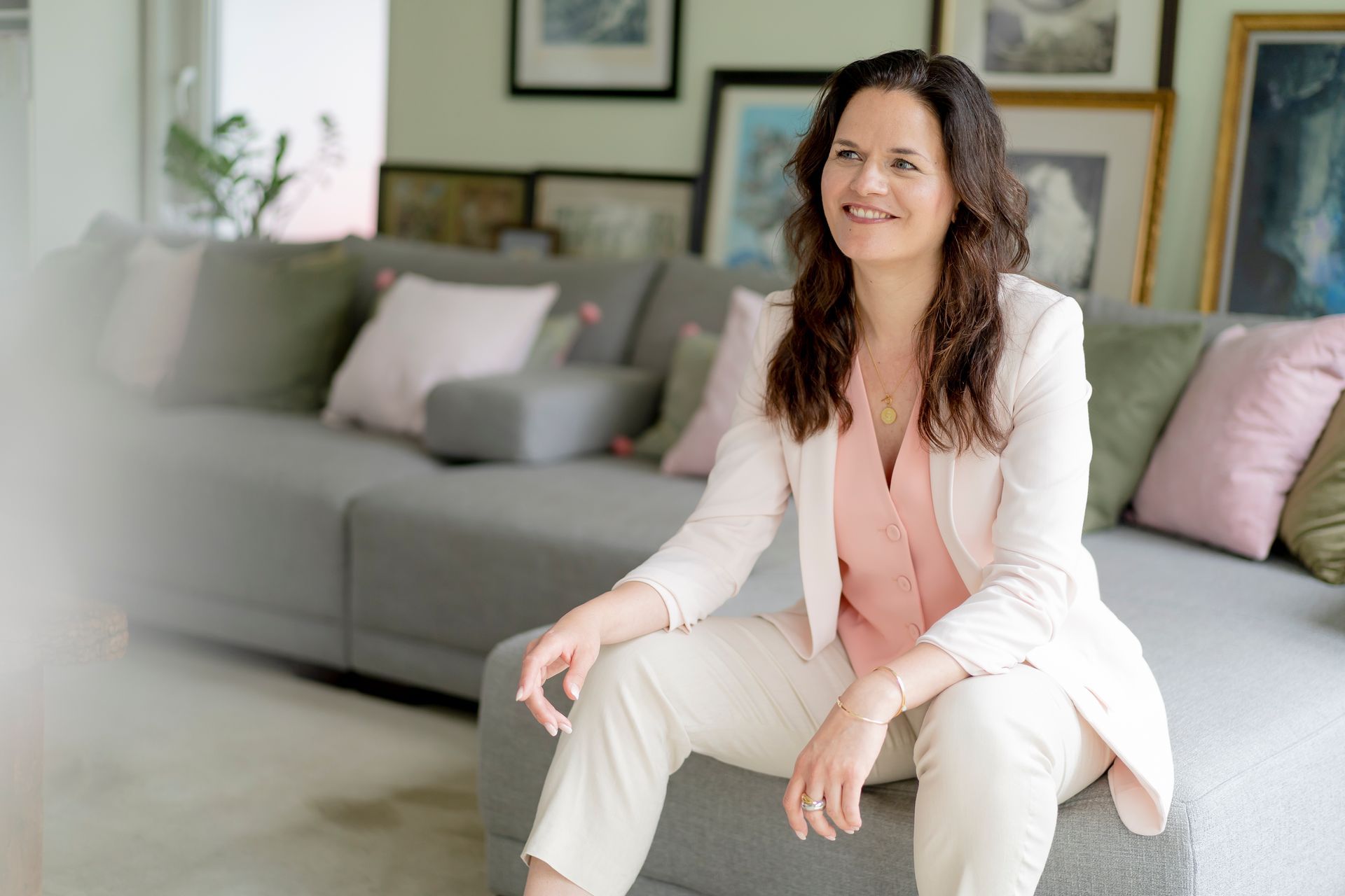 Traurednerin Kristina Kutz sitzt strahlend auf einem großen grauen Sofa. Sie trägt eine Kombi aus Blazer, Anzugweste, Hose.