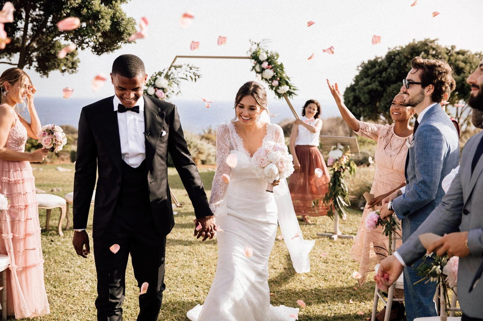 Ein Brautpaar läuft strahlend nach der Freien Trauung auf Mallorca durch seine Gäste hindurch. Im Hintergrund sieht man die Freie Traurednerin Kristina Kutz vor einem mit Blumen geschmückten Traubogen. Ganz in der Ferne ist das Mittelmeer zu sehen.