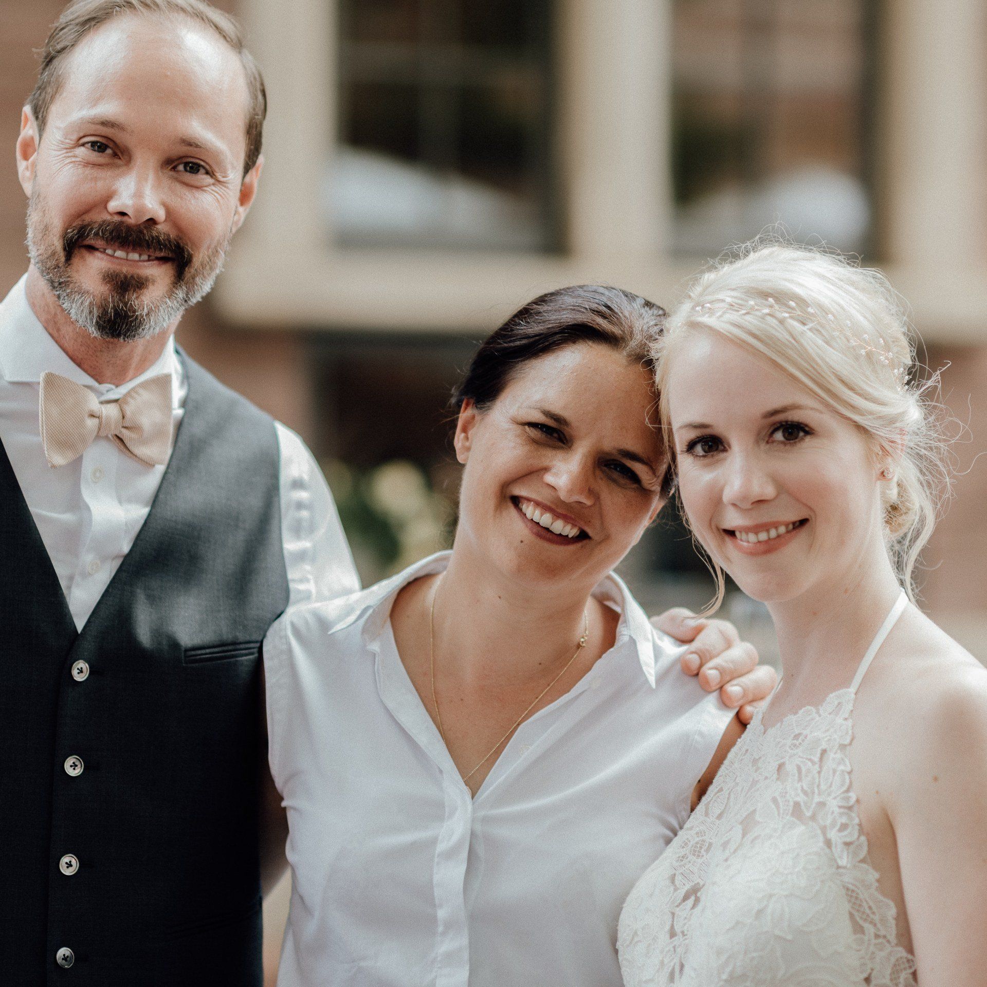 Die Traurednerin Kristina Kutz steht strahlend in der Mitte eines Brautpaares, das seine Arme um sie legt.