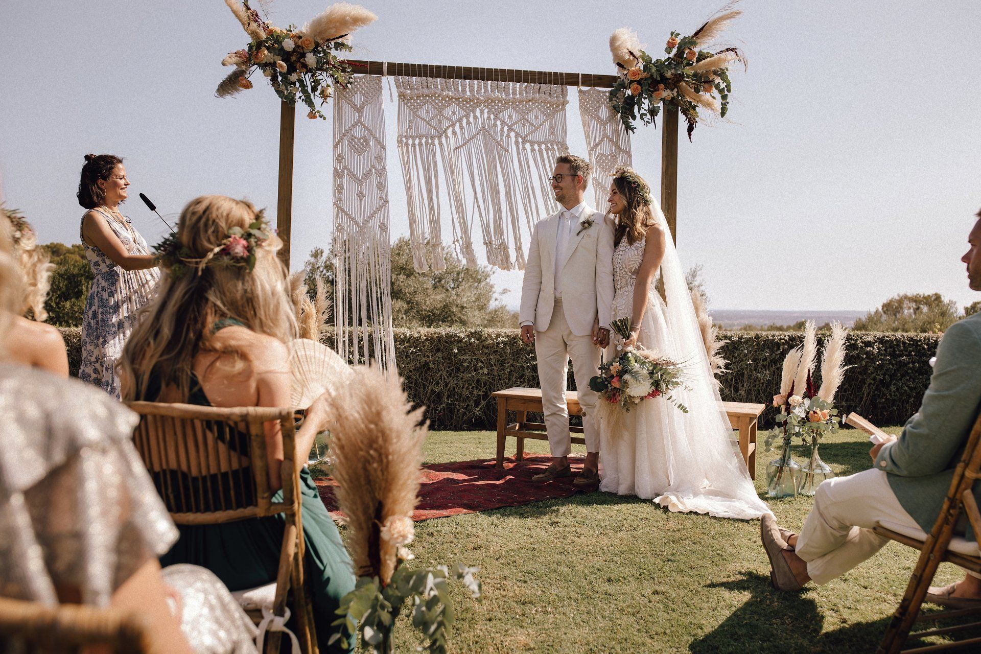Brautpaar steht vor einem mit Blumen verzierten Traubogen, strahlt Traurednerin während freien Trauung auf Mallorca an.