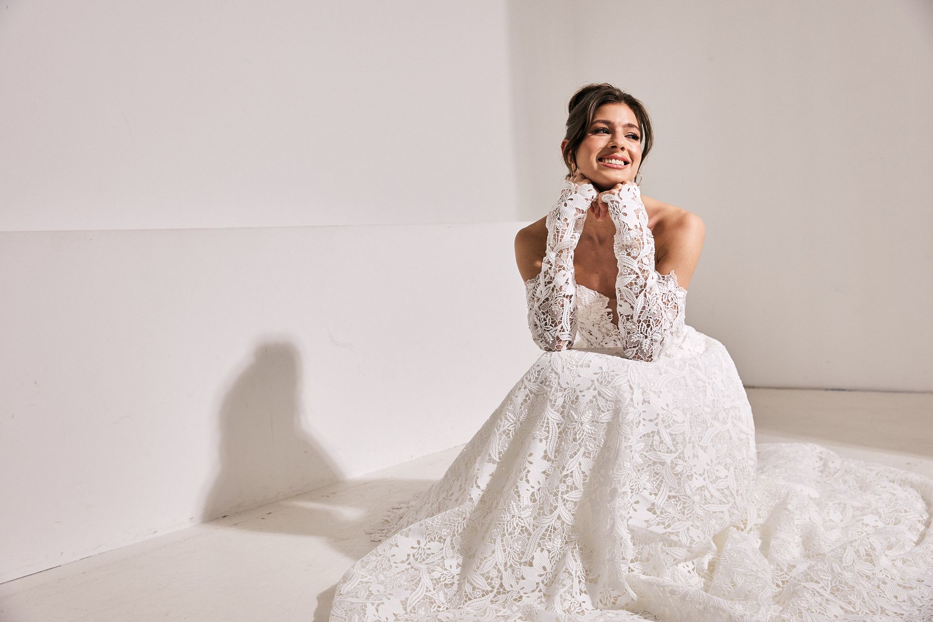 Braut hockt lächelnd in einem Brautkleid aus Spitze auf dem Boden.